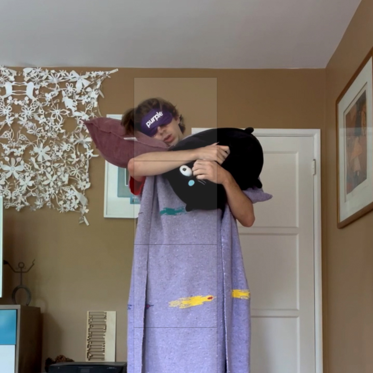 Man sleeping standing up, cuddling cat pillow while outfitted with a blanket and sleeping mask